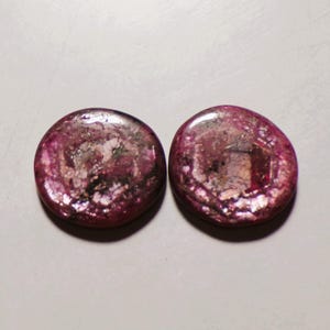 Puede incluir: Dos cabujones redondos y pulidos con una paleta de colores magenta intenso y rosa oscuro. Las piedras muestran una textura natural y moteada con inclusiones cristalinas visibles y superficies reflectantes que crean un aspecto brillante.