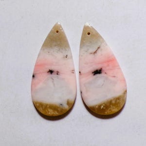 Peut inclure: Deux pendentifs en forme de goutte de pierre précieuse. Les pendentifs présentent un dégradé de couleurs blanc, rose et beige, avec de petites inclusions noires. Chaque pendentif a un trou percé en haut pour l'enfilage.