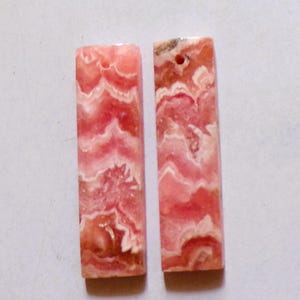 Puede incluir: Dos colgantes rectangulares de piedra preciosa de rodocrosita. Las piedras muestran una mezcla de patrones ondulados rosas, rojos y blancos. Cada colgante tiene un agujero perforado en la parte superior para ensartar. Las piedras preciosas están pulidas.