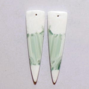 Op de afbeelding: Twee langwerpige, driehoekige keramische hangers met een gladde witte basis en verticale, penseelachtige strepen in licht saliegroen en donkerdere bosgroene accenten, elk voorzien van een klein rond gaatje aan de bovenzijde.