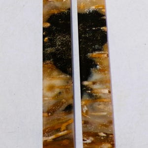 May include: Two rectangular, polished stone pendants. The pendants have a hole at the top for stringing. The stones feature a mix of colours, including black, brown, tan, and white, with a marbled appearance. The pendants are approximately 2.5 cm wide and 7.6 cm long.