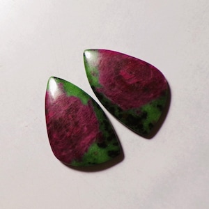 May include: Two polished, teardrop-shaped cabochons featuring a vibrant mix of deep magenta-red and forest green mineral inclusions. The stones have a smooth, reflective surface with distinct color patterns showing dark green and black speckles.