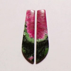 Puede incluir: Dos cabujones de piedra preciosa alargados y cónicos con una superficie pulida y un degradado de color distintivo. Las piedras muestran una sección superior de color magenta vibrante que transiciona hacia una base verde oscura y negra.