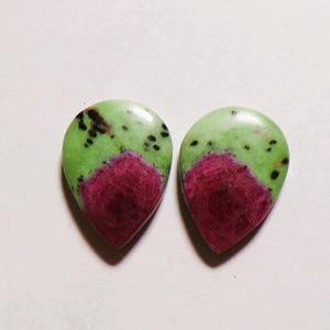 May include: Two teardrop-shaped cabochons featuring a smooth, polished surface. Each piece displays a vibrant lime green base colour with distinct, deep magenta-coloured inclusions and scattered small black spots across the upper green sections.