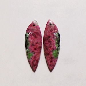 Peut inclure: Deux pendentifs en forme de marquise en pierre polie présentant un motif tacheté de magenta profond, de vert vif et d'inclusions noires. Chaque pièce possède un petit trou circulaire percé près de la pointe supérieure pour la suspension.