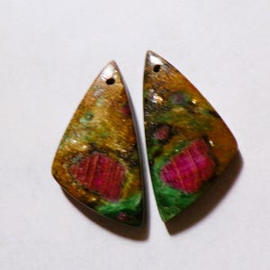 Peut inclure: Deux pendentifs en pierres précieuses de forme triangulaire. Les pendentifs présentent un mélange de couleurs, dont l'or, le vert et le rouge. Chaque pendentif a un trou en haut pour l'enfilage. Les pierres précieuses ont une finition polie.