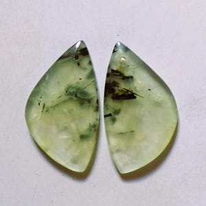Puede incluir: Dos cabujones de piedras preciosas de prehnita. Las piedras preciosas son de un verde pálido translúcido con inclusiones de color verde oscuro y negro. Cada piedra tiene forma de lágrima con un pequeño agujero perforado en la parte superior.
