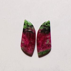 Puede incluir: Dos colgantes en forma de lágrima hechos de zoisita con inclusiones de rubí. Cada pieza presenta un centro magenta rosado vibrante, bordes verdes moteados y manchas minerales negras oscuras. Ambos colgantes tienen un pequeño orificio perforado.