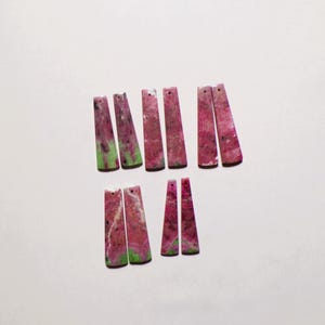 Op de afbeelding: Tien langwerpige, trapeziumvormige stenen hangers met een gevlekt patroon van levendig magenta, donkerroze en heldergroen. Elk stuk heeft een glad, gepolijst oppervlak en een klein, rond gaatje geboord nabij de bovenrand voor ophanging.