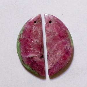 Peut inclure: Deux pendentifs en pierres précieuses semi-circulaires assortis. Chaque pendentif présente un mélange de teintes rose foncé et vertes, avec un trou percé en haut pour l'enfilage. La surface polie révèle des motifs naturels, suggérant un élément de bijouterie unique et artisanal.