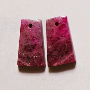 May include: Two rectangular, pink gemstone pendants with a hole drilled at the top for stringing. The pendants have a marbled appearance with darker pink and black veining throughout. The bottom edges are wider than the top edges.