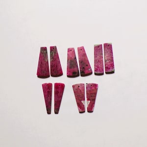 Puede incluir: Diez colgantes de piedra planos con forma trapezoidal dispuestos en cinco pares. Cada pieza presenta un patrón jaspeado en magenta vibrante y rosa oscuro con inclusiones gris oscuro y negras, e incluye un pequeño orificio perforado.