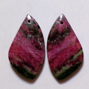 Peut inclure: Deux pendentifs en forme de goutte de pierre précieuse. Les pendentifs sont d'un magenta vibrant avec des inclusions vertes foncées et noires. Chaque pendentif a un petit trou en haut pour l'enfilage. Les pierres précieuses ont une surface polie et lisse.