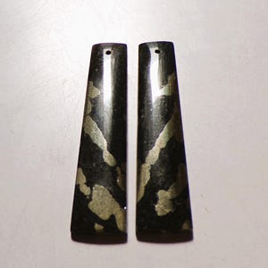 May include: Two elongated, tapered rectangular pendants made of polished black stone with irregular, metallic gold-coloured inclusions. Each piece features a small, circular hole drilled near the top edge for suspension, showcasing a smooth, reflective surface.