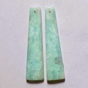 Op de afbeelding: Een paar lange, taps toelopende, platte hangers van licht muntgroene steen met subtiele witte en donkergroene gemarmerde patronen. Elk stuk heeft een klein, rond gaatje bij de bovenrand om op te hangen, met een gladde, gepolijste oppervlaktestructuur.