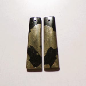 May include: Two rectangular pendants featuring a combination of black stone and metallic gold-toned pyrite inclusions. Each piece has a flat, elongated shape with a small, circular hole drilled near the top edge for suspension or attachment purposes.