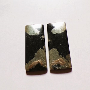 Puede incluir: Dos cabujones rectangulares con una superficie negra lisa y pulida con inclusiones metálicas irregulares de tono dorado. Las piezas tienen una forma plana y alargada con bordes superiores ligeramente redondeados y acabado brillante.
