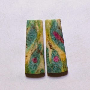 Pode incluir: Dois cabochões trapezoidais com um padrão marmoreado em verde-azulado, creme e amarelo com distintas inclusões circulares magenta. As pedras têm uma superfície lisa e polida e apresentam uma composição mineral natural e multicolorida.