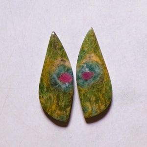 Op de afbeelding: Twee traanvormige cabochons met een gevlekt groen en geel oppervlak met een centrale blauwe halo rondom een levendige roze robijninsluiting. Deze gepolijste edelstenen vertonen natuurlijke kleurvariaties en organische patronen over het gehele oppervlak.