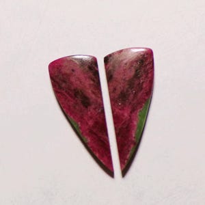 Puede incluir: Dos piedras preciosas de rubí zoisita pulidas en forma de lágrima, con un centro magenta rosado vibrante, inclusiones negras oscuras y un borde verde distintivo en los lados exteriores, mostrando una superficie lisa, reflectante y textura natural.