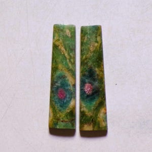 Peut inclure: Deux cabochons allongés de forme trapézoïdale présentant une surface marbrée verte et jaune avec un motif circulaire bleu sarcelle distinct centré autour d'une petite inclusion rose vif au milieu de chaque pièce de pierre.