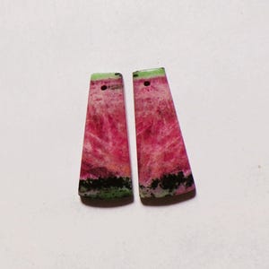 Può includere: Due pendenti in pietra trapezoidali caratterizzati da un centro rosa vibrante con bande verdi sui bordi superiore e inferiore. Ogni pezzo presenta un piccolo foro circolare praticato vicino alla parte superiore, con una texture cristallina.