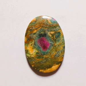 Könnte beinhalten: Ein ovaler Cabochon-Edelstein mit einer polierten Oberfläche, die ein wirbelndes Muster aus moosgrünen, goldgelben und dunkelbraunen Tönen aufweist, zentriert durch einen deutlichen, leuchtend magentafarbenen Einschluss als Blickfang.
