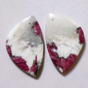 May include: Two gemstone pendants with a teardrop shape. The pendants are white with streaks of gray and deep pink. Each pendant has a small hole at the top for stringing.