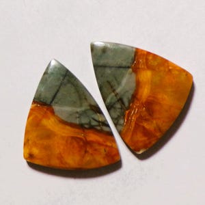 May include: Two triangular cabochons featuring a distinct color split. Each piece displays a smooth, polished surface with a combination of muted grey-green tones and vibrant, swirling orange patterns throughout the stone material.