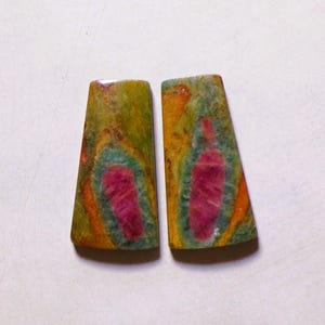 Op de afbeelding: Twee trapeziumvormige cabochons met een levendige mix van kleuren. Elke steen toont een centrale langwerpige magentakleurige insluiting omringd door een blauwgroene halo, tegen een gevlekte achtergrond van olijfgroen, mosterdgeel en oranje.