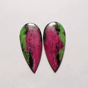 Puede incluir: Dos piedras preciosas de zoisita en forma de lágrima con un centro rosa magenta vibrante rodeado de inclusiones minerales verdes y negras moteadas. Estos cabujones pulidos muestran una superficie lisa y brillante con variaciones de color.