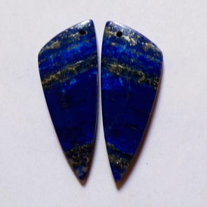May include: Two polished, teardrop-shaped lapis lazuli stones. The deep blue stones have gold and gray veining, and a small hole at the top for stringing. The stones are set against a white background.