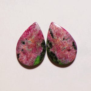 Puede incluir: Dos cabujones en forma de lágrima que presentan una mezcla vibrante de inclusiones minerales en magenta, rosa y verde oscuro. Las piedras pulidas muestran una textura cristalina moteada con variaciones de color distintas.