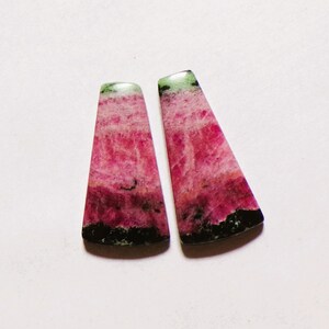 May include: Two trapezoidal cabochons featuring a vibrant mix of magenta, deep pink, and black mineral inclusions. Each stone displays a smooth, polished surface with distinct colour banding, transitioning from a dark base to a lighter top.