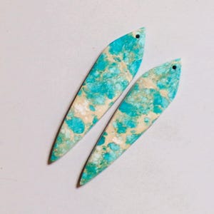 Op de afbeelding: Twee langwerpige, bladvormige hangers met een gemarmerd patroon van helder turkooisblauw en crèmewit. Elk plat, glad stuk heeft een klein gaatje geboord bij het puntige uiteinde aan de bovenkant voor bevestiging, met natuurlijke texturen.