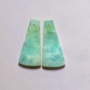 Op de afbeelding: Twee platte, trapeziumvormige hangers van lichtgroene en turquoise steen met een gemarmerd, doorschijnend uiterlijk. Elk stuk heeft een klein, rond gaatje in het midden bovenaan voor bevestiging, met zichtbare natuurlijke mineraalpatronen.