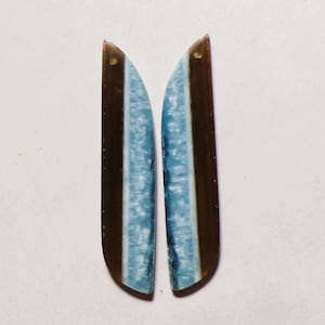 Puede incluir: Dos componentes de joyería alargados y curvos con una división de color vertical. Un lado es de color marrón oscuro translúcido, mientras que el otro es de color azul claro, texturizado y veteado. Cada pieza tiene un pequeño orificio circular arriba.