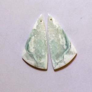 Puede incluir: Dos colgantes triangulares de piedra pulida. Cada colgante tiene un agujero en la parte superior para colgar. Las piedras presentan una mezcla de patrones arremolinados blancos, verde pálido y verde más oscuro, creando un efecto veteado natural. Los colgantes son probablemente para la fabricación de joyas.