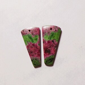 Puede incluir: Dos colgantes en forma de cuña hechos de zoisita con inclusiones de rubí, con una superficie pulida con patrones moteados de color rosa magenta vibrante y verde intenso. Cada pieza tiene un pequeño orificio circular cerca del borde.