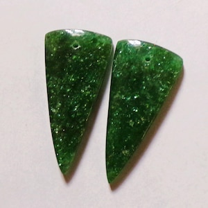 May include: Two triangular, dark green gemstone pendants with a speckled, glittery appearance. Each pendant has a small hole at the top for stringing. The gemstones are set against a white background.