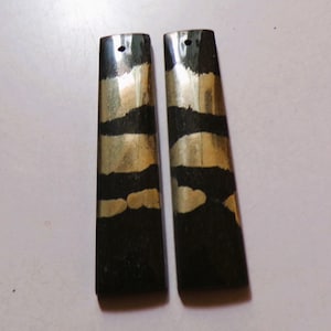 Puede incluir: Dos pendientes rectangulares con base negra y rayas horizontales doradas. Cada pendiente tiene un pequeño agujero en la parte superior para colgar. Los pendientes se muestran sobre una superficie clara.