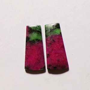 May include: Two trapezoidal ruby zoisite gemstones featuring a vibrant magenta pink base color with contrasting patches of bright green and dark black inclusions. These polished stones have a smooth, flat surface and a distinct tapered shape.