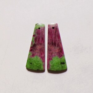 Pode incluir: Dois pingentes trapezoidais de pedra polida com uma mistura vibrante de cores magenta e verde brilhante. Cada peça tem uma superfície lisa e plana, com um pequeno orifício circular perfurado perto da borda superior para pendurar.