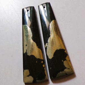 Puede incluir: Dos colgantes alargados en forma de trapecio. Los colgantes son negros y dorados con un agujero en la parte superior para ensartar. El oro tiene un brillo metálico y el negro tiene un acabado mate. Los colgantes son probablemente para hacer joyas.