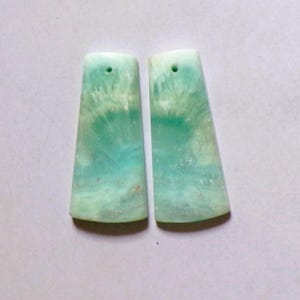 May include: Two trapezoidal pendants made of polished light blue and white stone. Each piece features a smooth, marbled texture with a distinct radial pattern and a small, circular hole drilled near the top edge for suspension in jewelry making.
