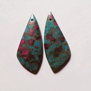 Könnte beinhalten: Zwei tropfenförmige Edelsteinanhänger. Die Anhänger haben einen türkisfarbenen Hintergrund mit unregelmäßigen Flecken in Dunkelrosa und Braun. Jeder Anhänger hat oben ein kleines Loch zum Auffädeln.