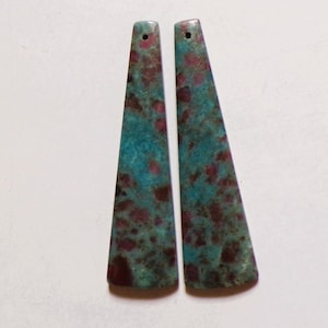 May include: Two gemstone pendants with a trapezoid shape. The pendants are teal and burgundy, with a hole drilled at the top for stringing. The pendants are likely for jewelry making.