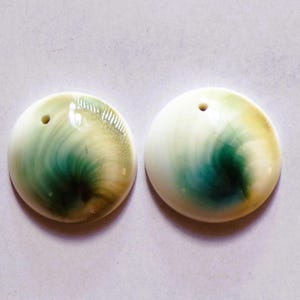 May include: Two round, polished shell pendants with a hole drilled at the top. The pendants have a swirling pattern of green, yellow, and white. These pendants are ideal for jewelry making.