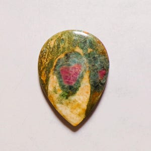 Puede incluir: Un cabujón de piedra preciosa pulida en forma de lágrima que presenta una superficie moteada con remolinos de color verde oliva, crema y bronceado, centrado con una distintiva inclusión de color rosa magenta vibrante y un acabado liso y brillante.