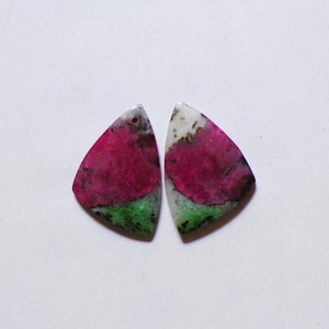 Op de afbeelding: Twee driehoekige cabochons met een levendige combinatie van diep magenta, heldergroene en crèmekleurige mineraalpatronen. Elk stuk heeft een glad, gepolijst oppervlak met een klein voorgeboord gaatje bij de bovenste hoek.